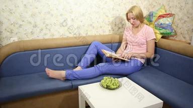 金发女郎坐在沙发上，看书，吃美味的葡萄