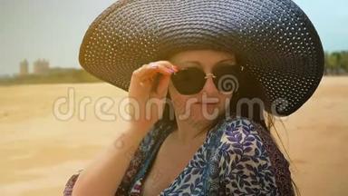 美丽的快乐女孩戴着蓝色的<strong>帽子</strong>和眼镜，坐在阳光明媚的<strong>夏日</strong>海滩上。 近距离射击。 慢动作