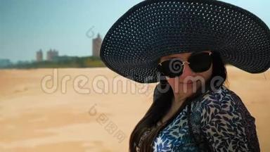美丽的快乐女孩戴着蓝色的帽子和眼镜，坐在阳光明媚的夏日海滩上。 近距离射击。 慢动作