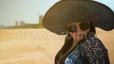 美丽的快乐女孩戴着蓝色的帽子和眼镜，坐在阳光明媚的夏日海滩上。 近距离射击。 慢动作
