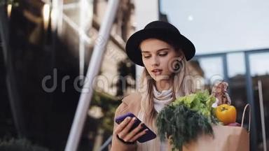 年轻漂亮的女人穿着时髦的外套，走在街上，拿着<strong>一包</strong>产品，用智能手机