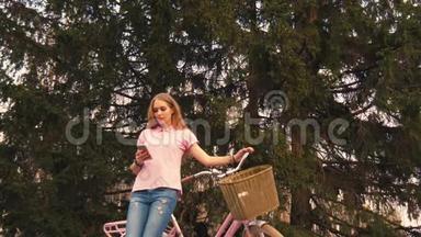 姜红头女孩坐在自行车上，在夏季公园的智能手机上发短信。 穿着粉色衬衫和牛仔裤