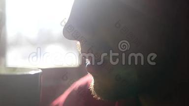 清晨，一个留着胡子的男人喝着浓墨重彩的热咖啡，坐在窗前晒太阳，动作缓慢