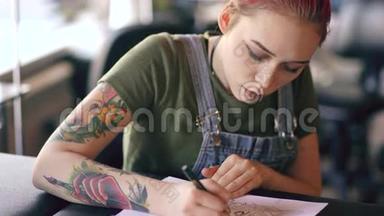 特写年轻迷人的红发女纹身艺术家坐在桌子旁，为工作室的纹身创作素描。