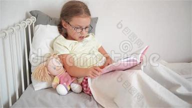 戴眼镜的小女孩躺在床上翻书。 也许孩子生病了。 她旁边坐着一个洋娃娃。
