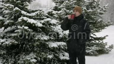 一个年轻人在冬天的森林里喝<strong>热</strong>水瓶里的<strong>热饮料</strong>。 大雪纷飞。 那个人在雪地里环顾四周