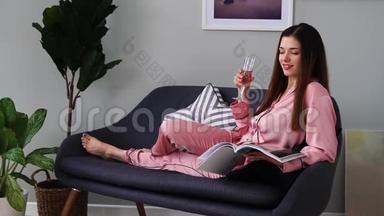 一个穿着粉红色睡衣的漂亮女孩看着一本光滑的杂志躺在沙发上，从<strong>玻璃杯</strong>里喝<strong>香槟</strong>。 在