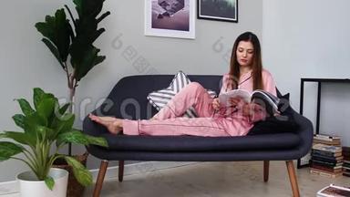 一个穿着粉红色睡衣的漂亮女孩看着一本光滑的杂志躺在沙发上，从玻璃杯里喝香槟。 在