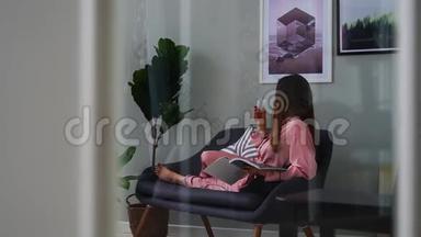 一个穿着粉红色睡衣的漂亮女孩看着一本光滑的杂志躺在沙发上，从玻璃杯里喝香槟。