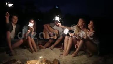 在日落黄昏时，朋友们在海滩上闪闪发光地庆祝。 年轻男女围坐在篝火旁