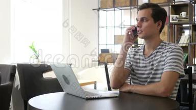 男人在手机上说话的肖像，坐在办公桌前，看着笔记本电脑