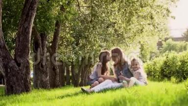 妈妈和两个女儿坐在苹果树附近的草地上。 日落时公园里的孩子和母亲。 投标书
