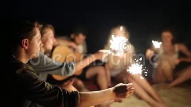 在日落黄昏时，朋友们在海滩上闪闪发光地庆祝。 年轻男女围坐在篝火旁