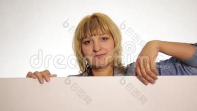 微笑的幸福女人站在后面，靠在一个白色的空白广告牌或标语牌上，拇指指向顶部，躲藏起来