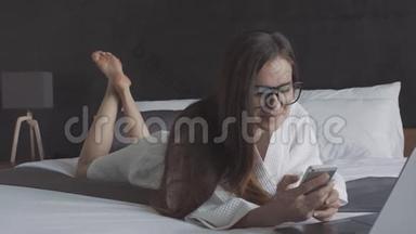 穿着<strong>白色浴袍</strong>和戴着智能手机和笔记本电脑的眼镜的女人躺在床上微笑。 在线商务。