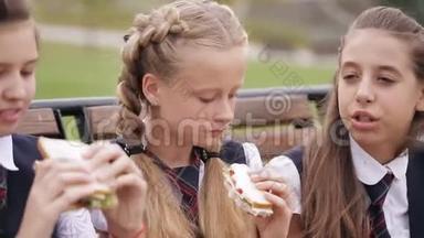 女大学生穿着同样的校服，坐在花园里的长凳上吃三明治，笑着。