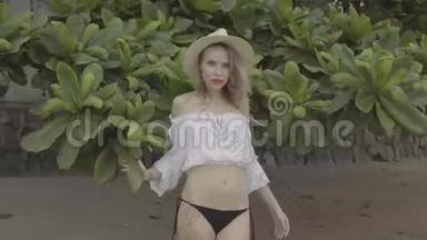 美丽的女人在夏日穿着波霍风格的时尚，穿着白色衬衫，比基尼和帽子，微笑着，在海滩上摆姿势。