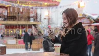 美丽的年轻女子在<strong>旋转</strong>木马前的圣诞集市上，使用智能<strong>手机</strong>，喝着热茶