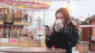 美丽的年轻女子在旋转木马前的圣诞集市上，使用智能手机，喝着热茶