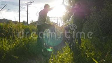 两个男朋友互相<strong>击掌</strong>，在日落时骑自行车穿过草地。 朋友骑自行车