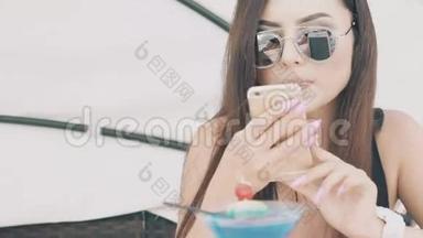 穿着泳衣的漂亮女孩在沙滩桌上用手机配鸡尾酒