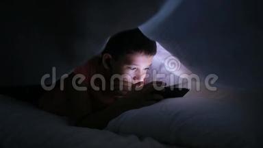 在床上玩手机或智能手机的男孩。 躲在毯子下面