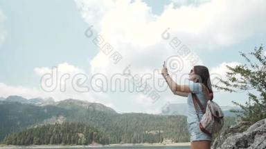 年轻女子用智能手机在山湖前拍照。 美丽的白种人女孩在一个女孩子中度过的时光