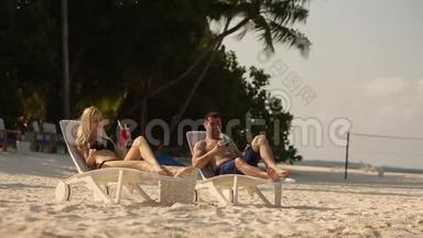 年轻的家庭夫妇在热带海滩晒日光浴和喝彩色鸡尾酒。 在异国度假的蜜月中情人