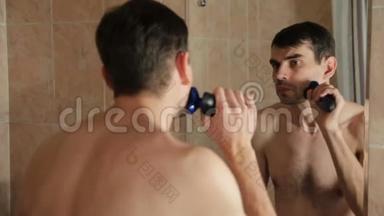 年轻人在浴室里用电动剃须刀刮胡子。 他在浴室里用剃刀刮胡子，看着里面