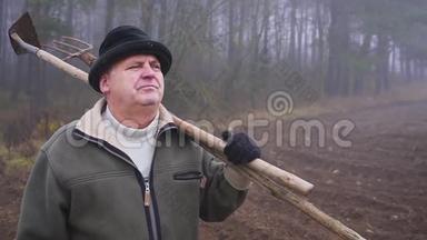戴着帽子的高级农民，手里拿着一把泥泞的锄头和田野上的草叉。 体力劳动。 福克
