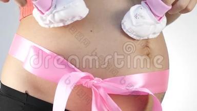 穿着内衣的怀孕妈妈在玩婴儿鞋。 腹部有粉红色的丝带。 慢动作