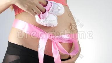穿着内衣的怀孕妈妈在玩婴儿鞋。 腹部有粉红色的丝带。 慢动作