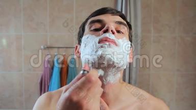 一个人用剃刀在泡沫中剃了他的脸。 他在镜子前刮胡子