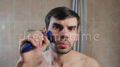 年轻人在浴室里用电动剃须刀刮胡子。 他在浴室里用剃刀刮胡子，看着里面