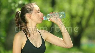 苗条的年轻女人在训练后喝水。 女运动员休息，她喝水，在炎热的跑步中外出