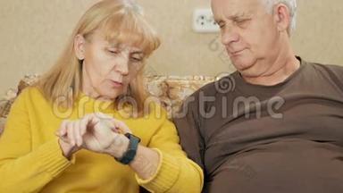 年长夫妇在家里的沙发上看智能手表上的信息。 他们仔细阅读并阅读重要的文章