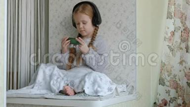 戴着耳机<strong>辫子</strong>的快乐<strong>小女孩</strong>看着手机视频，坐在窗台上的一个房间里，藏了一条毯子
