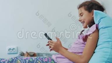 可爱的女孩和狗宠物躺在床上的智能手机肖像。 小女孩和狗宠物玩网络游戏