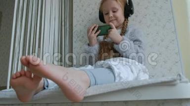 戴着耳机<strong>辫子</strong>的快乐<strong>小女孩</strong>看着手机视频，坐在窗台上的一个房间里，藏了一条毯子