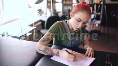 年轻迷人的红头发妇女纹身艺术家坐在桌子上，为室内工作室纹身创作素描。