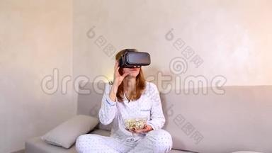 女孩看视频在VR眼镜与爆米花坐在沙发在明亮的卧室晚上。