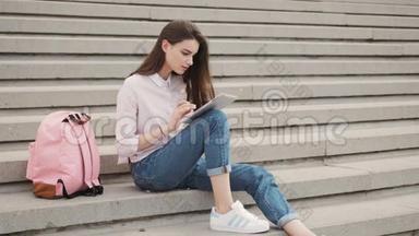 年轻漂亮的时尚女孩坐在台阶上，双手握在平板电脑上。 女生在社交网络中交流