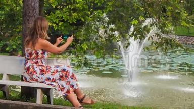 金发女孩坐在长凳上用Iphone拍喷泉