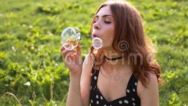 女孩吹肥皂泡。 年轻漂亮的女人坐在公园的草地上玩泡泡。 慢速视频