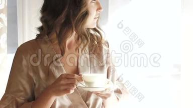穿睡衣的漂亮女孩在窗户附近喝了一杯咖啡。 慢慢地