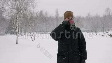 一个年轻人在冬天的森林里喝<strong>热</strong>水瓶里的<strong>热饮料</strong>。 大雪纷飞。 那个人看着镜头