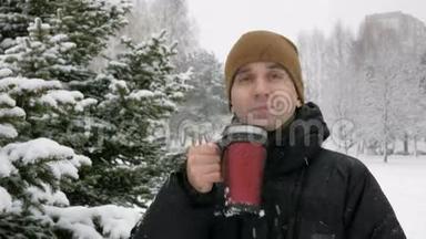 一个年轻人在冬天的森林里<strong>喝热水</strong>瓶里的热饮料。 大雪纷飞。 那个人看着镜头