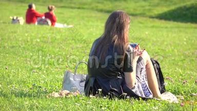 美丽的黑发女孩穿着黑色连衣裙，在草坪上使用她的智能手机。 休闲时间。 4K视频