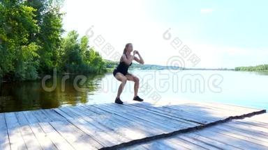 美丽的，运动的年轻金发女人伸展，做不同的运动，跳跃，跳跃，蹲。 湖泊，河流，蓝色