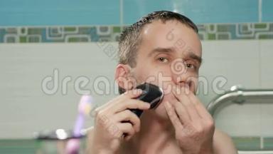 年轻迷人的男人在浴室里用电动剃须<strong>刀刮胡子</strong>。 他看着镜子里的照相机。 视察